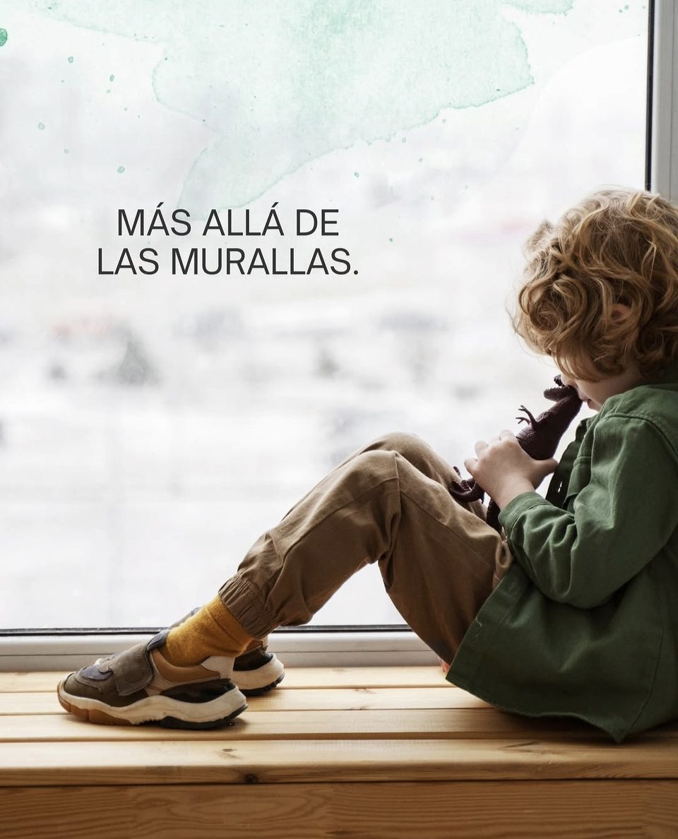 Un niño de espaldas, sentado junto a una ventana, abraza un dinosaurio de juguete; sobre el vidrio, el texto «Más allá de las murallas»